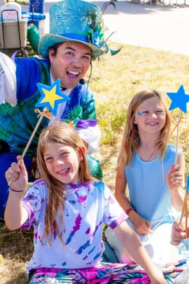 A Happily Ever Laughter performer and two smiling children hold star wands together outdoors during a recurring entertainment program.