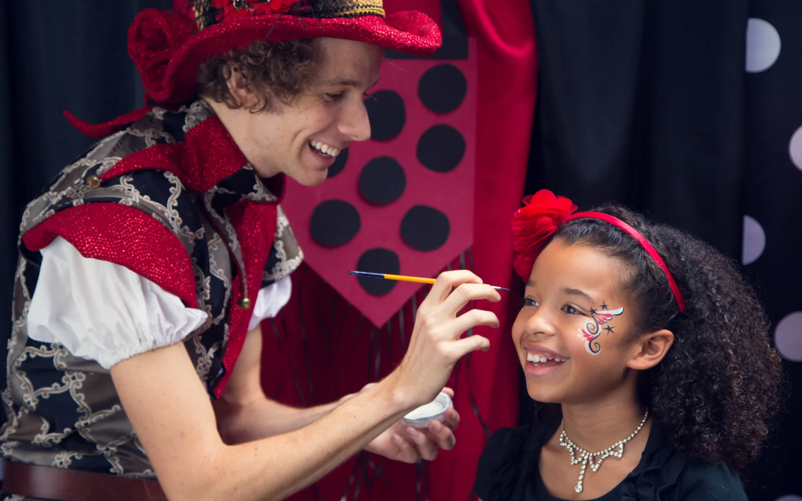 A Happily Ever Laughter face painter in a red costume painting a smiling child’s face at a party.