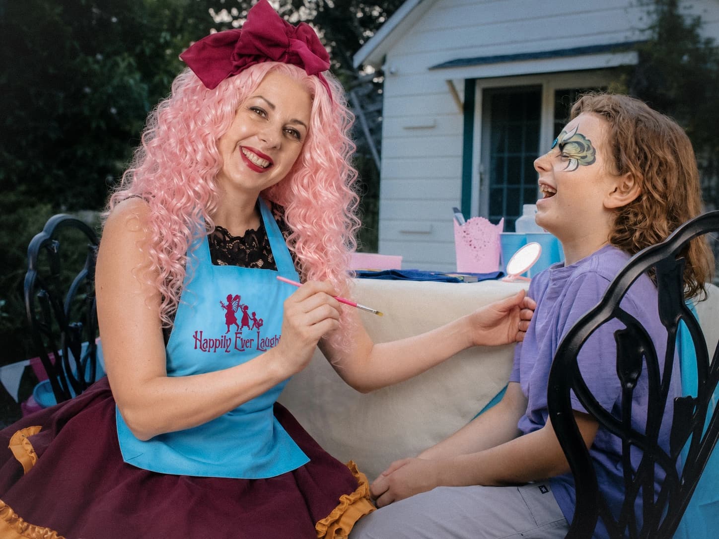 A Happily Ever Laughter face painter with pink hair paints a design on a smiling child at an outdoor party while other children look on.