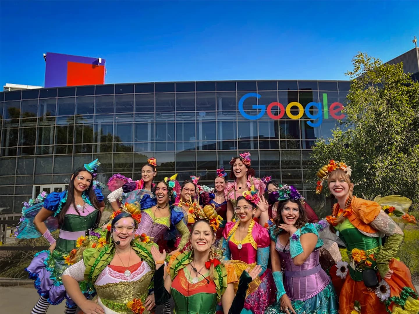 The Happily Ever Laughter team of faerie performers in colorful costumes posing together in front of the Google building.