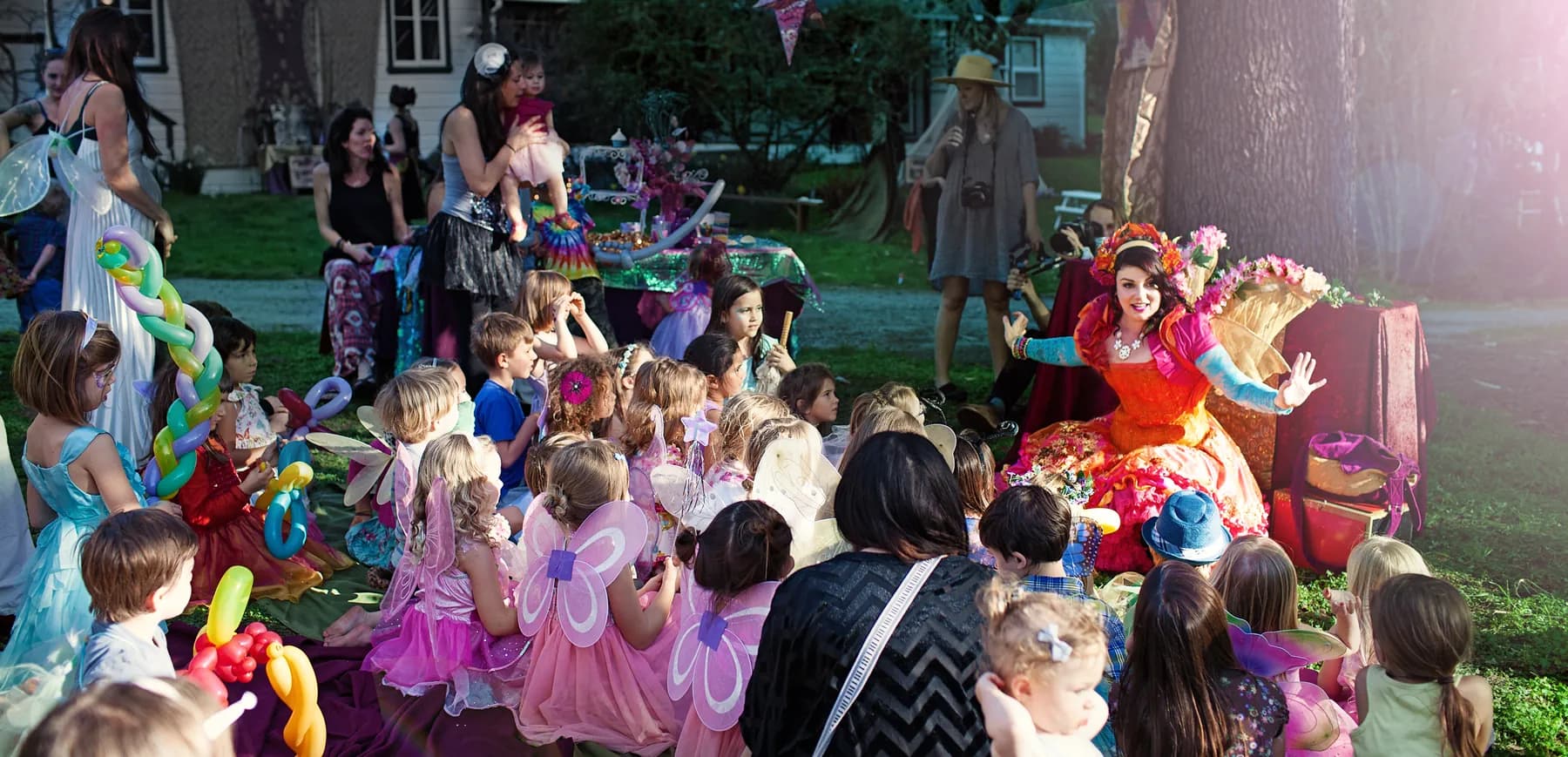 A HELP Faerie in a flower crown greeting a large group of delighted children at an outdoor garden party.