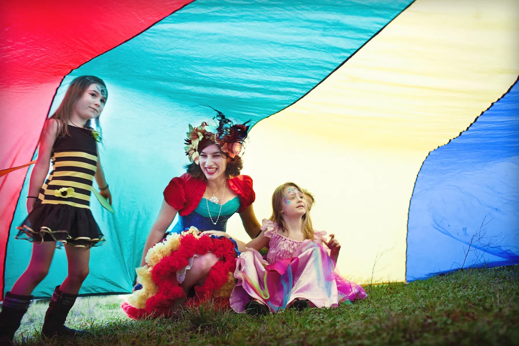 A HELP Faerie playing with two costumed children beneath a billowing rainbow parachute on the grass.