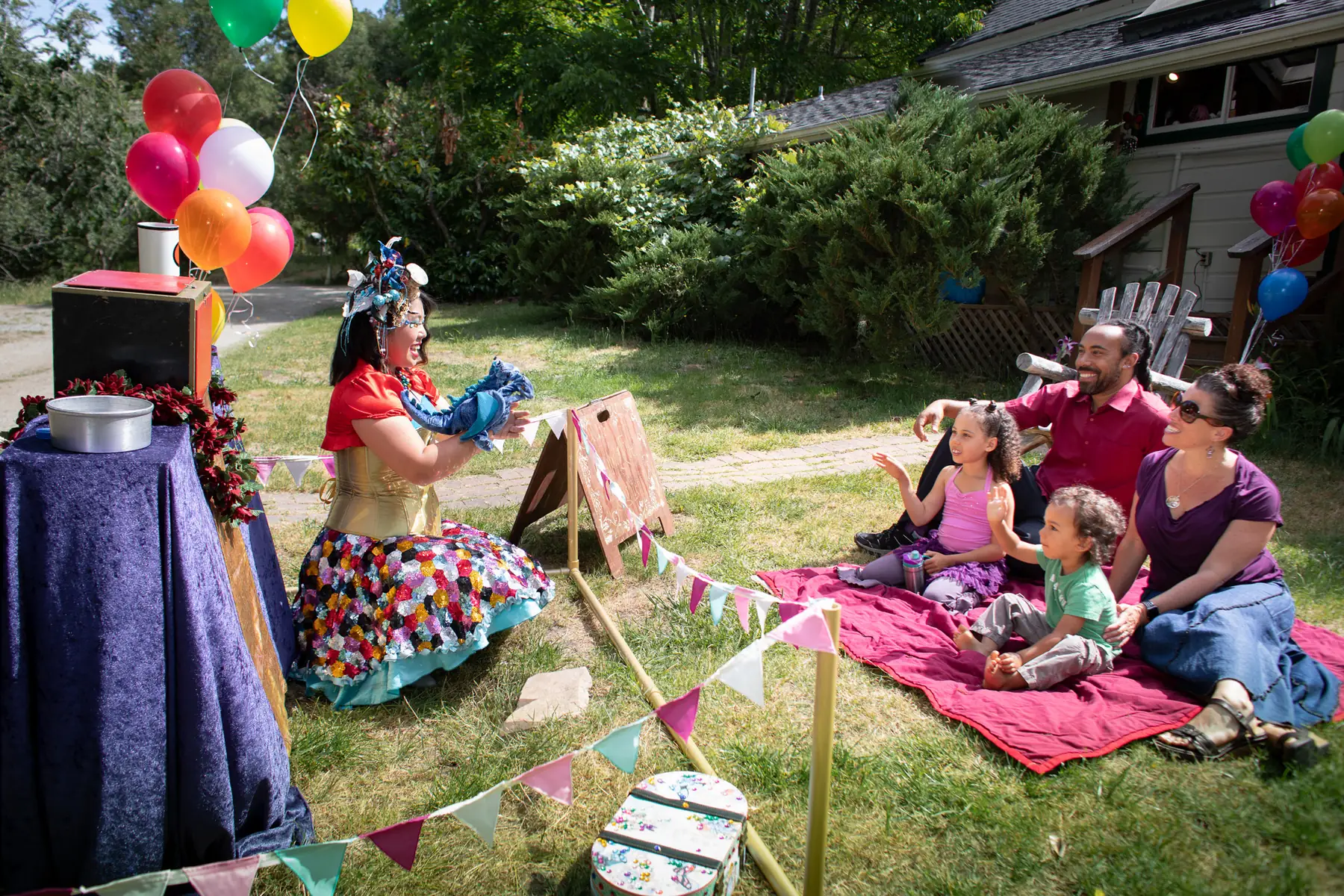 A HELP Faerie leading a puppet activity for a diverse family seated together on a picnic blanket.