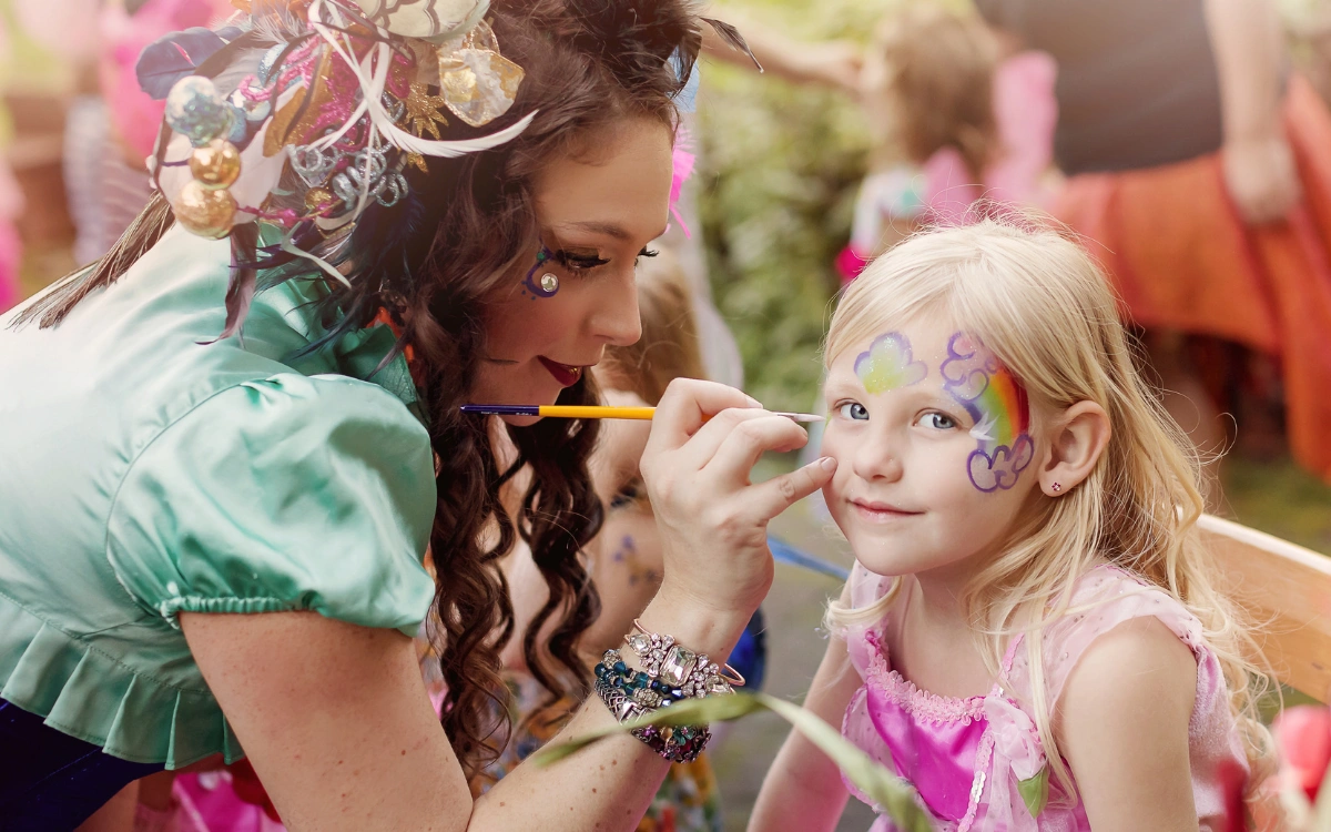 A Happily Ever Laughter face painter applying a glittery design to a child's face outdoors.