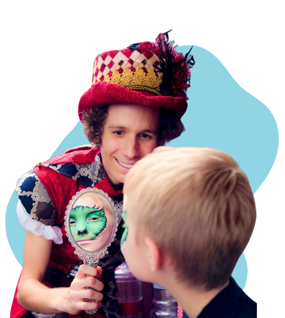 A Happily Ever Laughter face painter in a jester costume holding a hand mirror that shows a child’s freshly painted green-monster design.