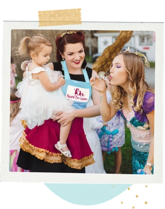 A Happily Ever Laughter faerie holding a toddler while painting another child’s face at an outdoor party.